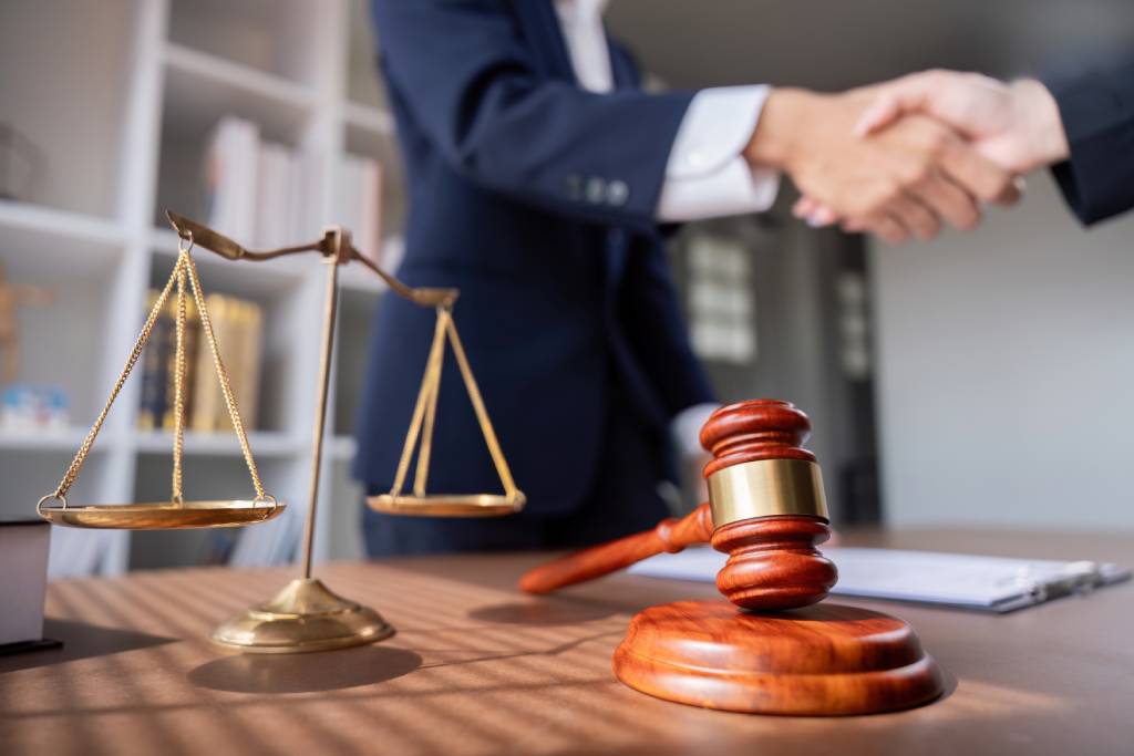 Two lawyers shaking hands over a desk with scales and a gavel.